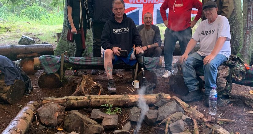 A group of people camping in a forest in front of a log fire with a kettle